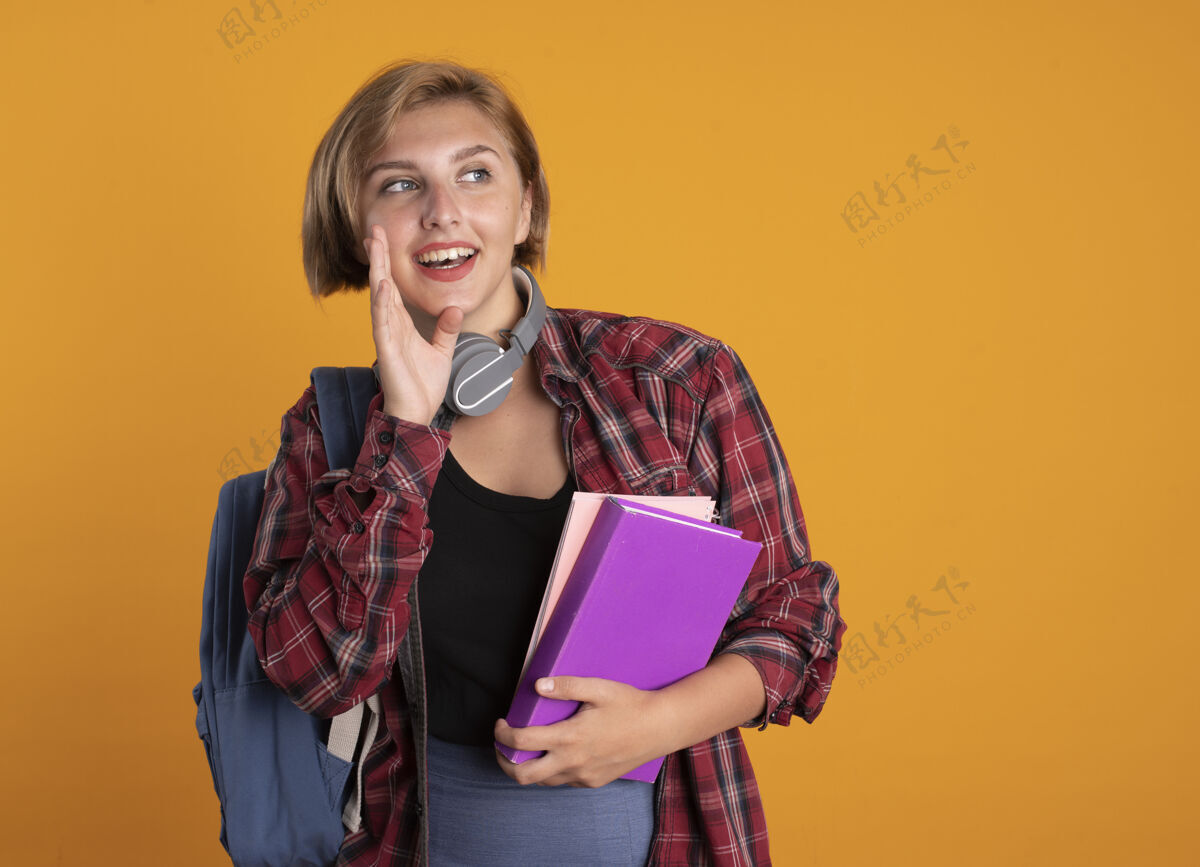 穿快乐的年轻斯拉夫女学生戴着耳机 背着背包 手贴着嘴 手里拿着书和笔记本持有欢乐橙色