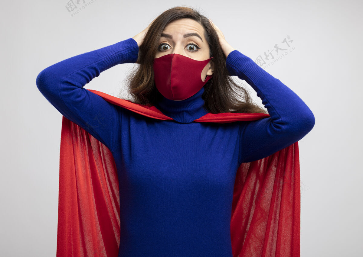 穿震惊的白种人超级英雄穿着红色斗篷戴着红色防护面具的女孩把手放在头上看着镜头保护头超级英雄