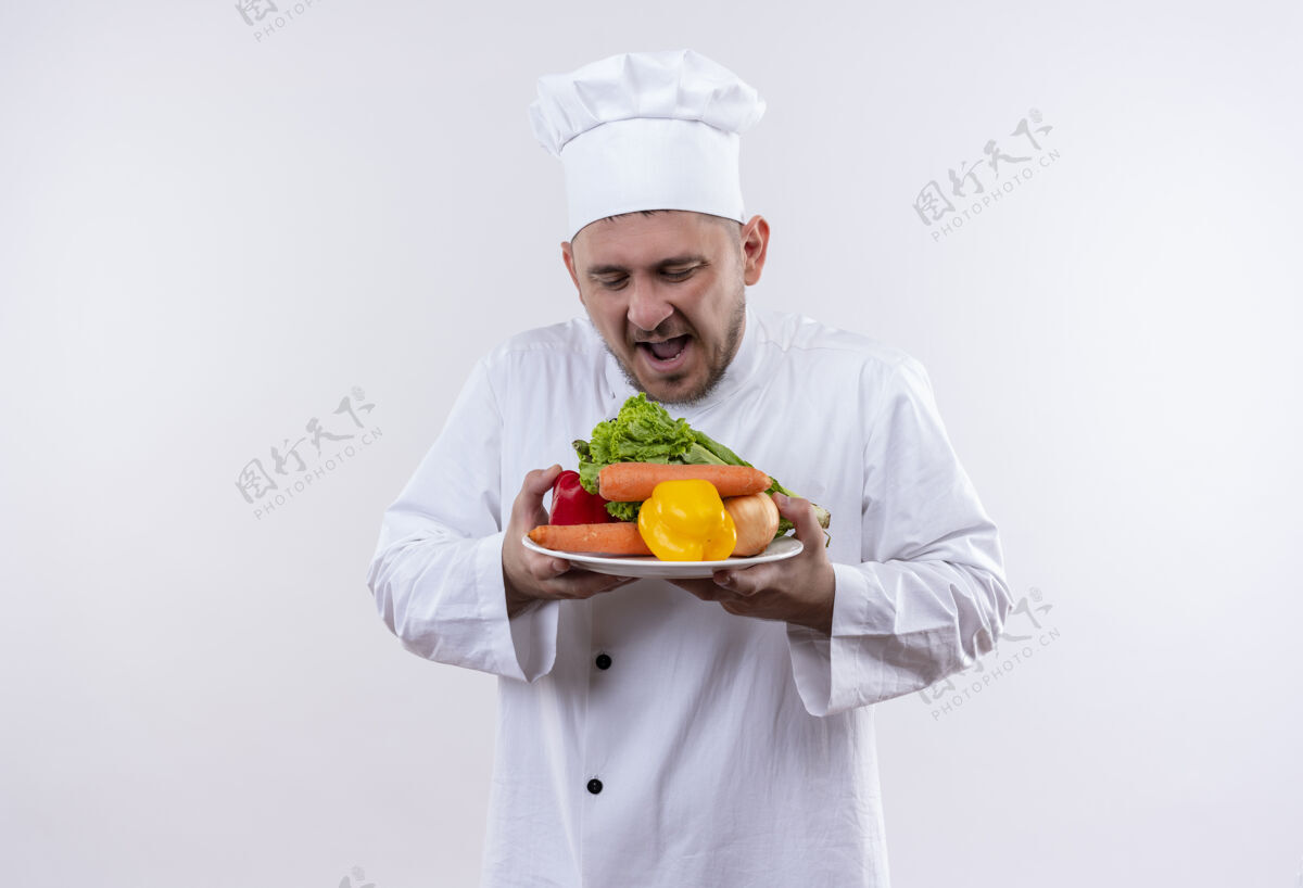 制服印象深刻的年轻帅哥厨师在厨师制服拿着蔬菜盘看着他们在孤立的白墙上手持年轻蔬菜
