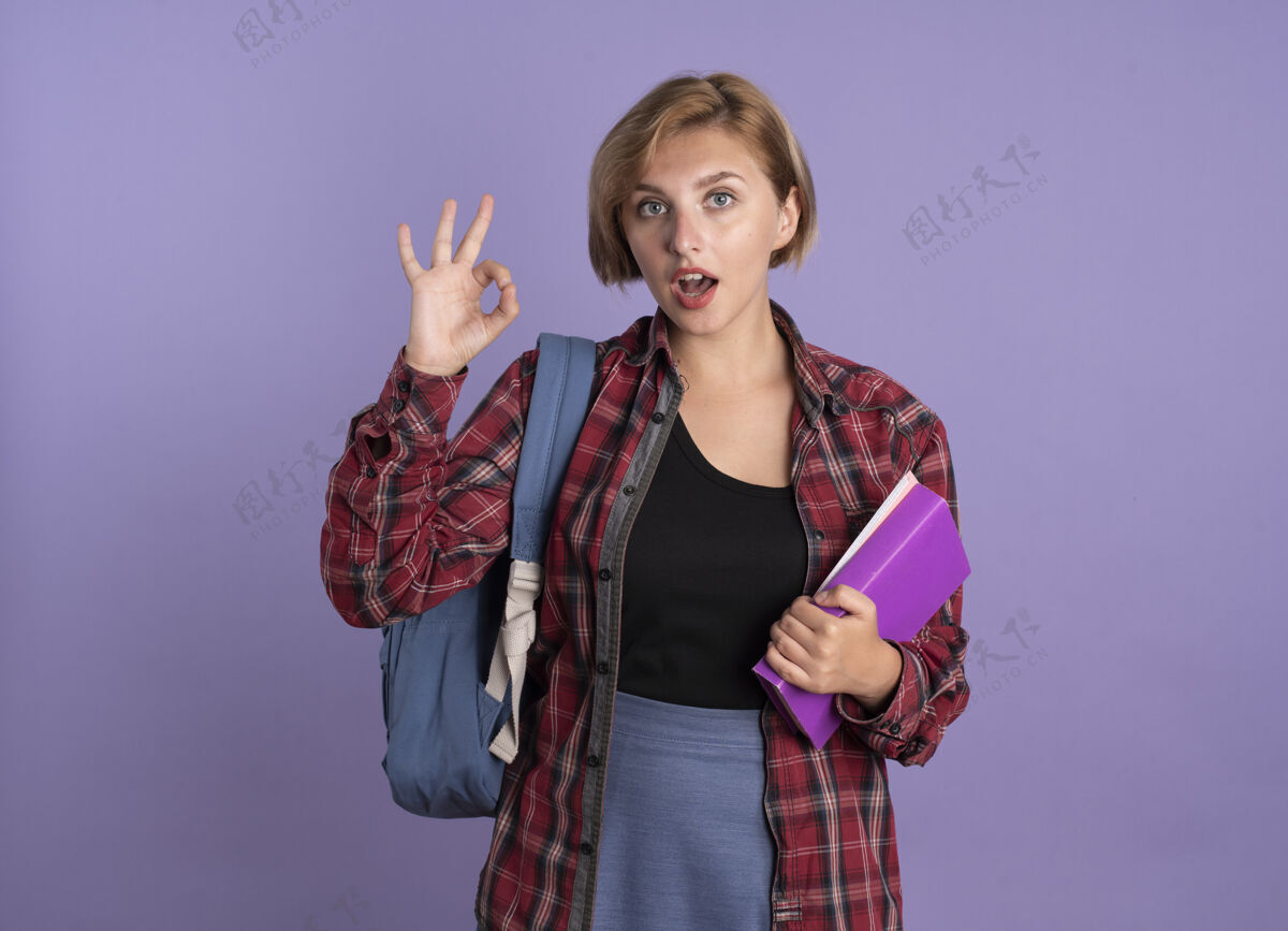 标志惊讶的年轻斯拉夫学生女孩背着背包拿着书和笔记本手势行手势背包空间学生