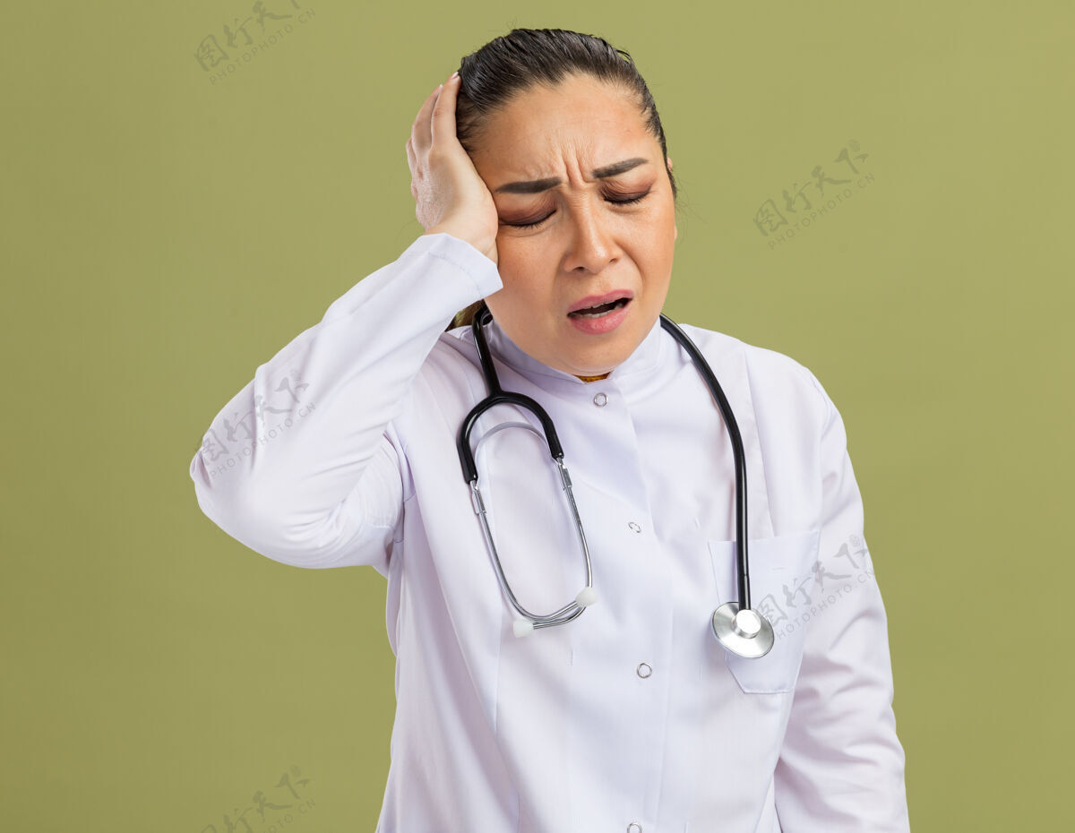 医生年轻的女医生 穿着白药衣 脖子上戴着听诊器 看着不舒服 站在绿墙上摸着头痛的头头女人药