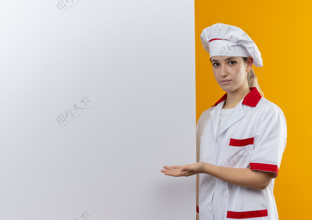 信心自信的年轻漂亮厨师穿着厨师制服站在旁边 用手指着隔离在橙色墙上的白色墙壁 还有复制空间近手年轻