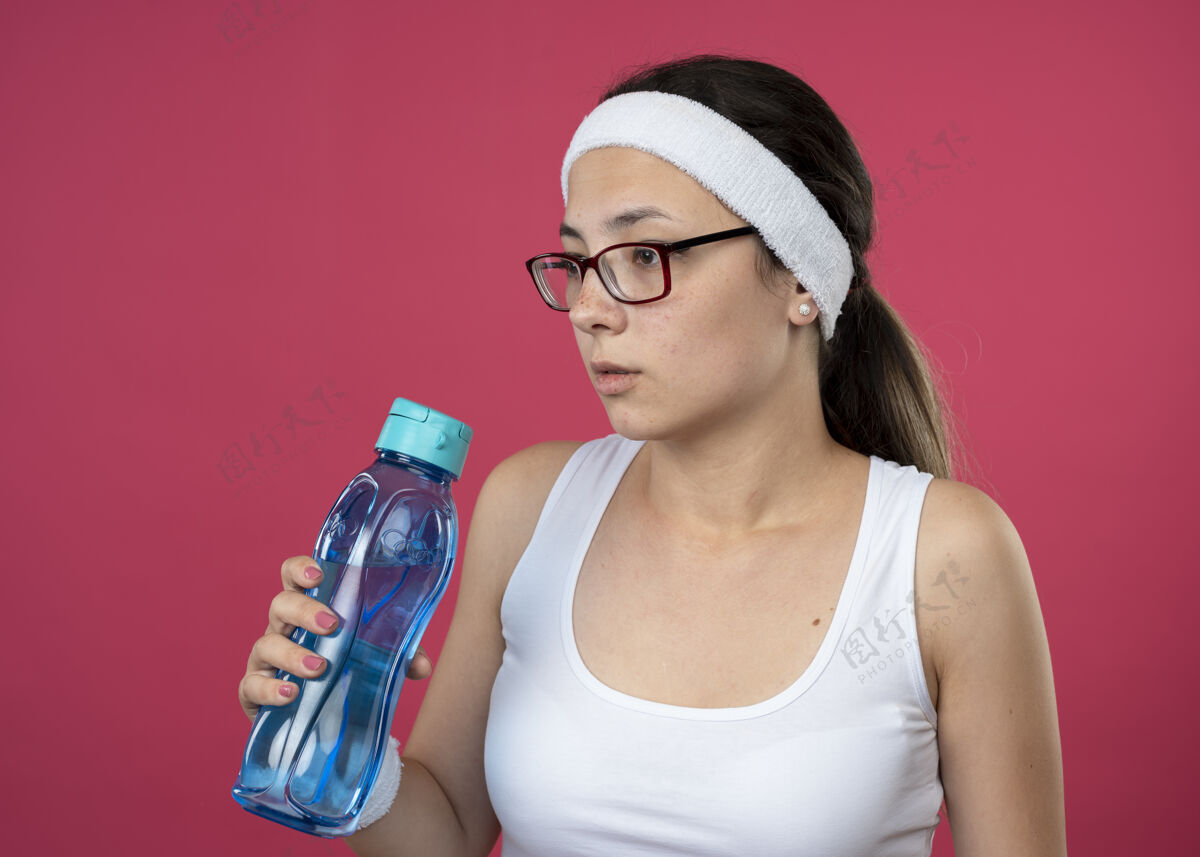 焦虑焦急的年轻运动女孩戴着眼镜 戴着头带和腕带 拿着水瓶 看着旁边瓶子女孩粉色