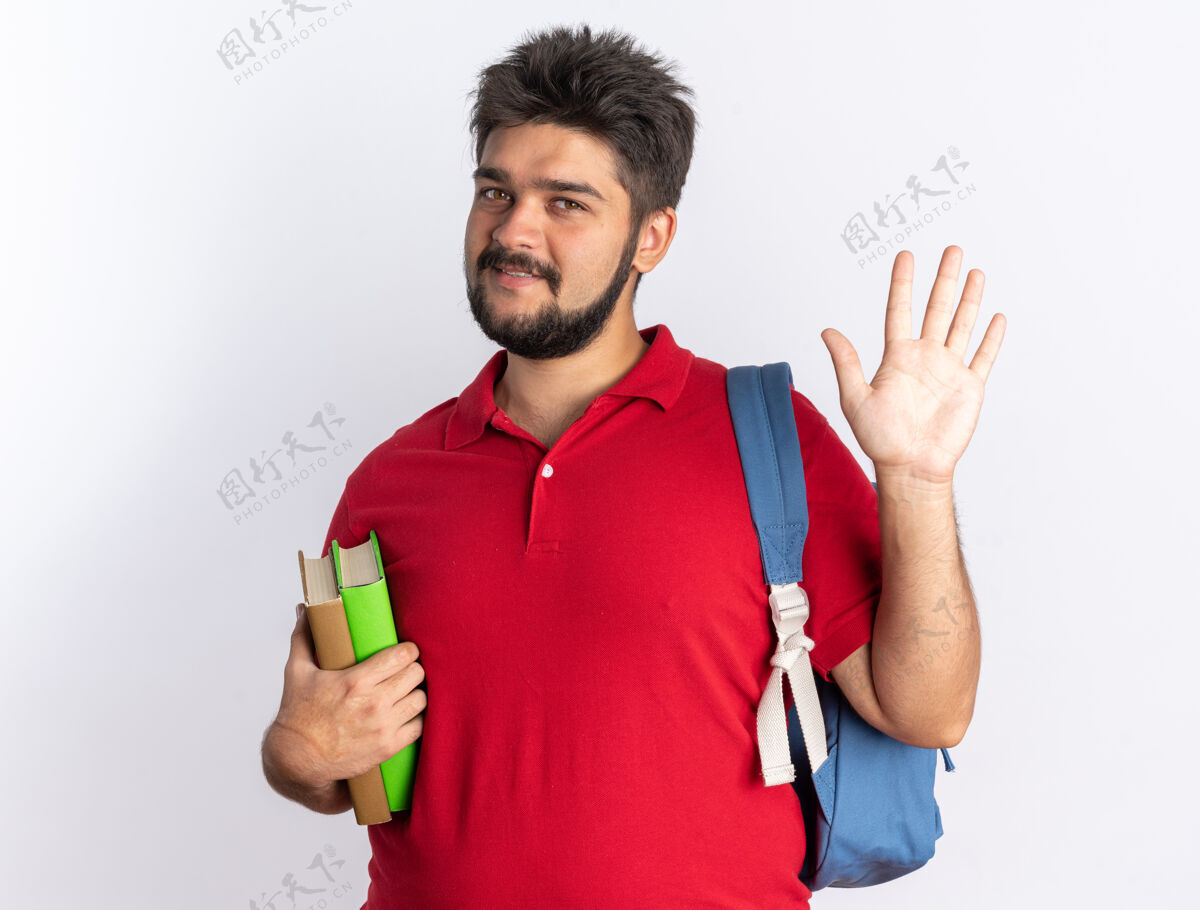 手年轻的留着胡子的学生 穿着红色马球衫 背着背包 拿着笔记本 面带微笑 站着挥手挥手学生红色