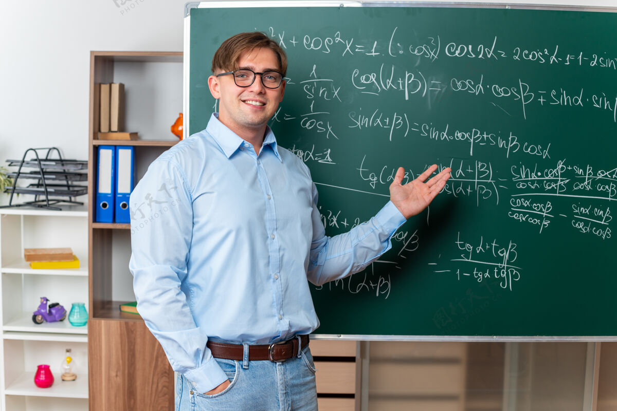 积极戴着眼镜的年轻男老师站在教室里数学公式的黑板旁愉快而积极地讲解功课快乐年轻教室
