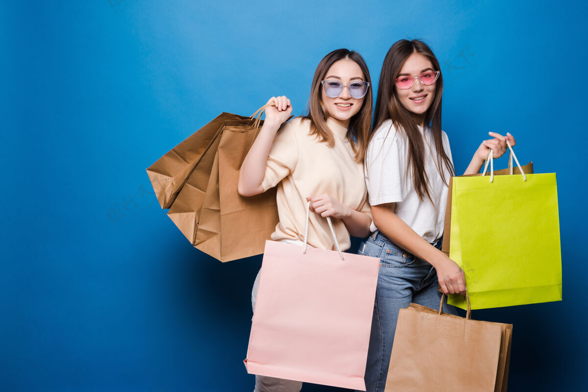 女人快乐的两个女人 蓝色的墙上挂着五颜六色的购物袋衣服一起商场
