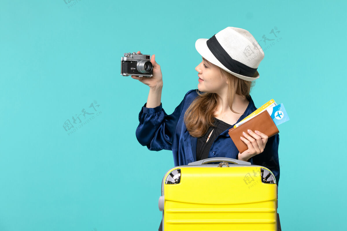 年轻女子前视图：手持相机和机票的年轻女子乘坐蓝色背景的女子海上旅行飞机机票旅行帽子