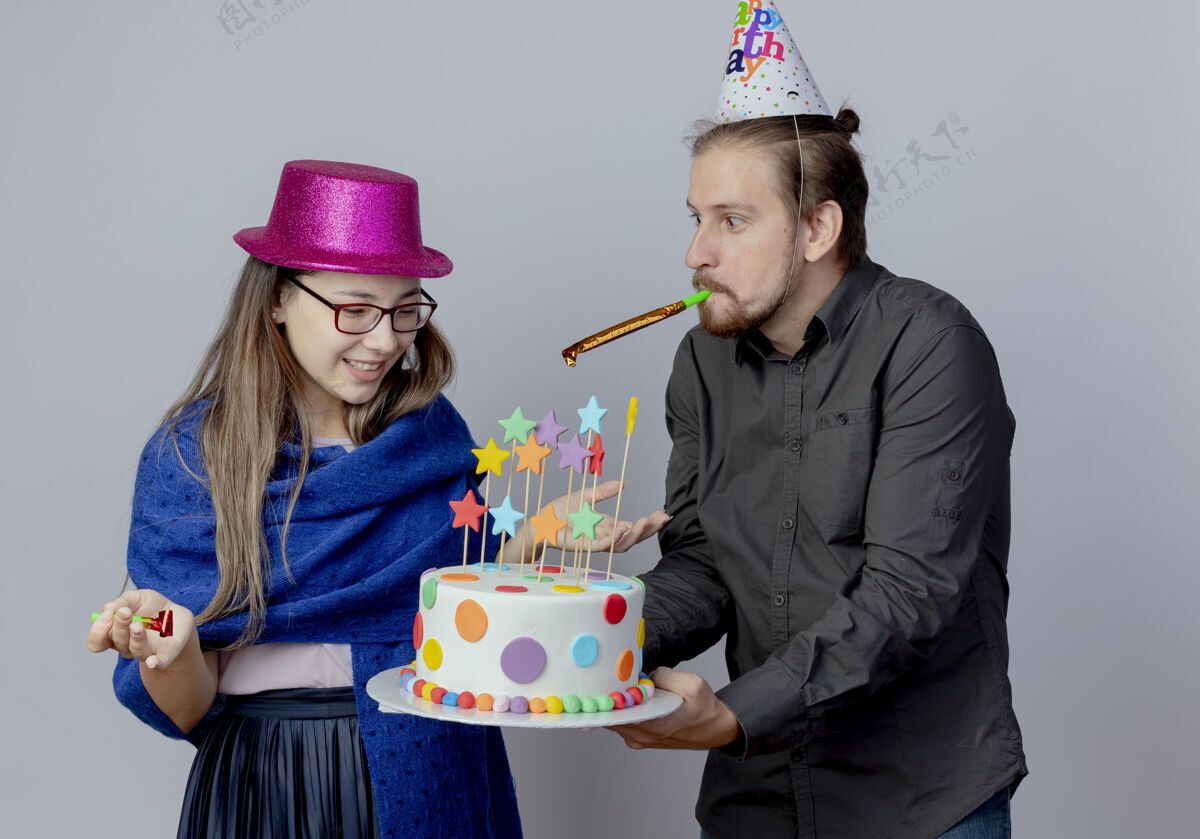 女孩快乐的帅哥戴着生日帽拿着蛋糕吹着口哨看着惊讶的戴着粉色帽子戴着眼镜的年轻女孩举着口哨看着蛋糕惊喜帽子空间