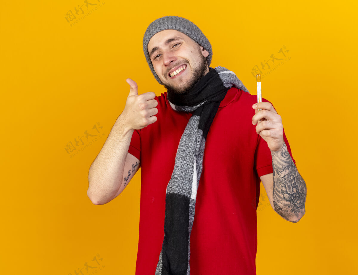疾病微笑着的年轻白种人 戴着冬天的帽子和围巾 竖起大拇指拿着温度计向上冬天年轻人