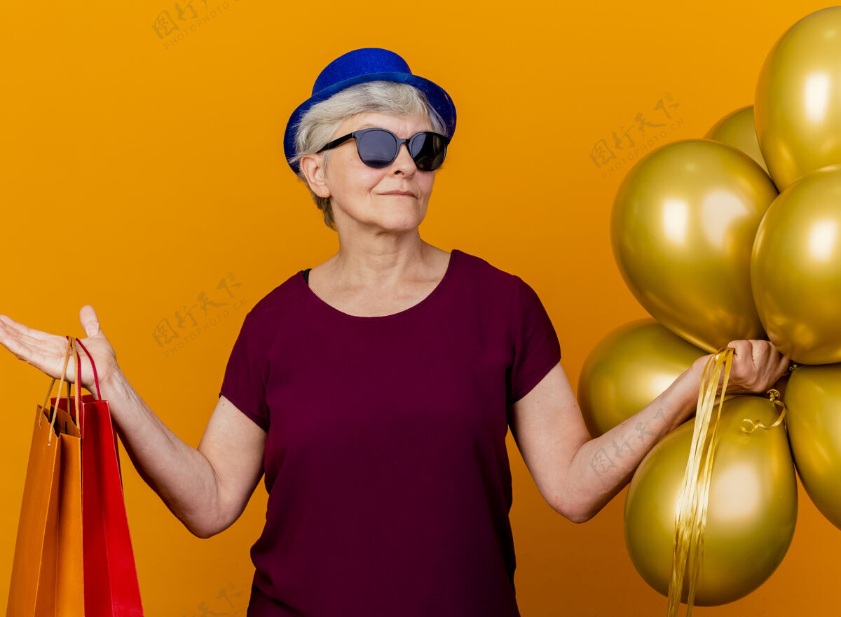 纸戴着派对帽 戴着太阳眼镜 手里拿着氦气球和纸购物袋的自信的老妇人老人包太阳