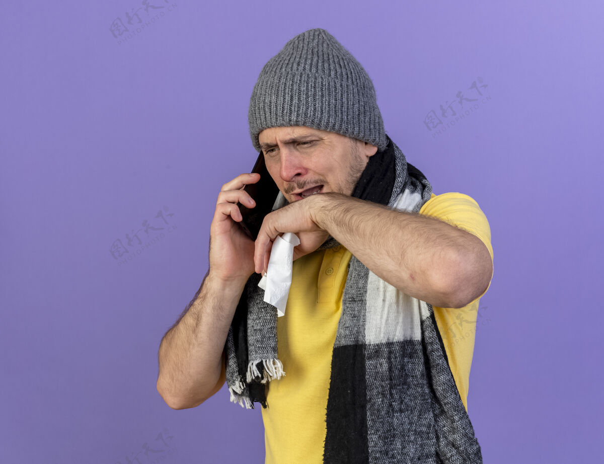 男人恼怒的年轻金发病斯拉夫男子戴着冬季帽子和围巾拿着纸巾讲电话斯拉夫人年轻人疾病