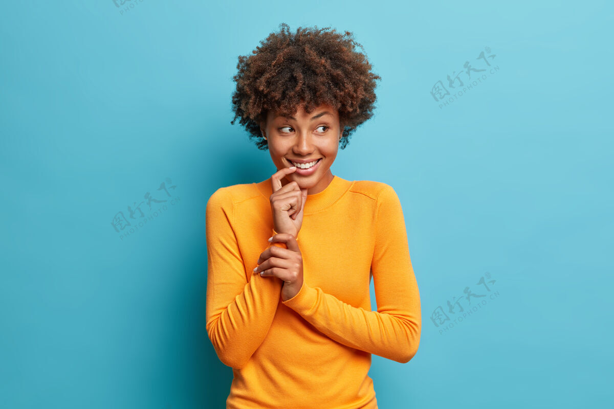 人类漂亮开朗的非裔美国女人微笑着温柔地穿着休闲长袖套头衫看起来很快乐地站在蓝色的墙上摆姿势思考室内快乐