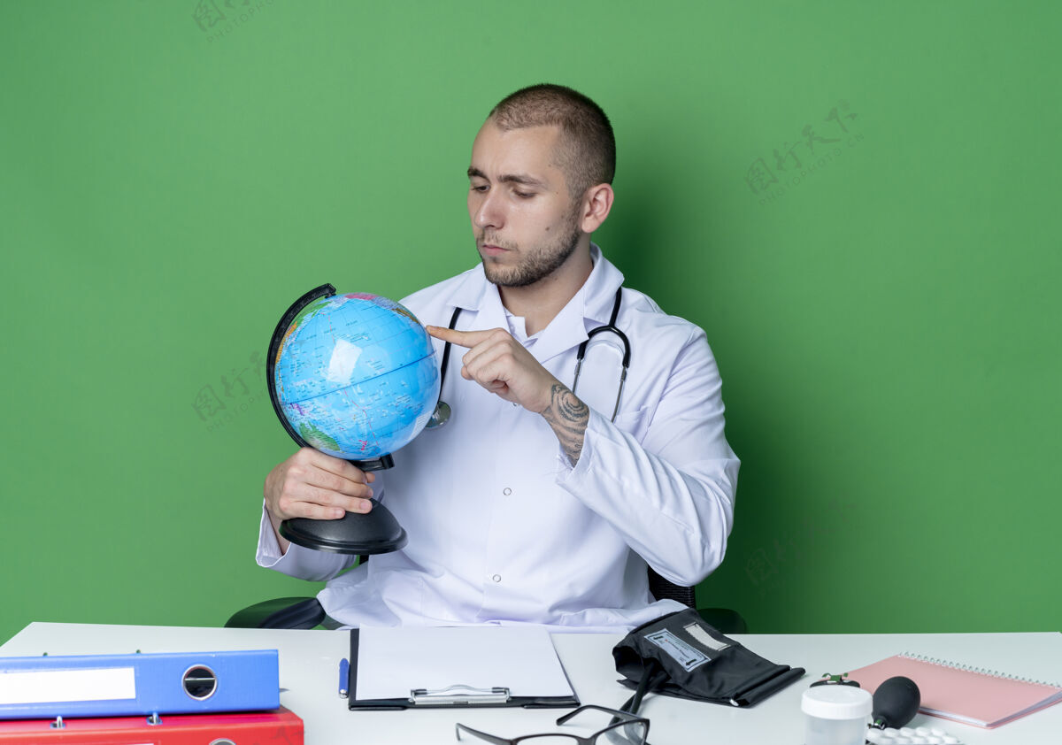 坐着体贴的年轻男性医生 穿着医用长袍 听诊器 坐在办公桌旁 拿着工作工具 看着地球仪 把手指放在隔离在绿色墙上的地球仪上绿色表情地球仪