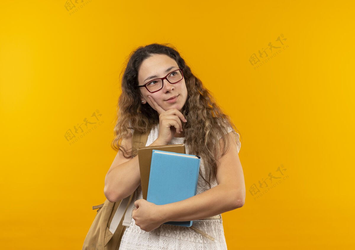 下巴体贴的年轻漂亮的女学生戴着眼镜 背着书包 手放在下巴上 看着隔离在黄色墙上的一面壁板脸年轻