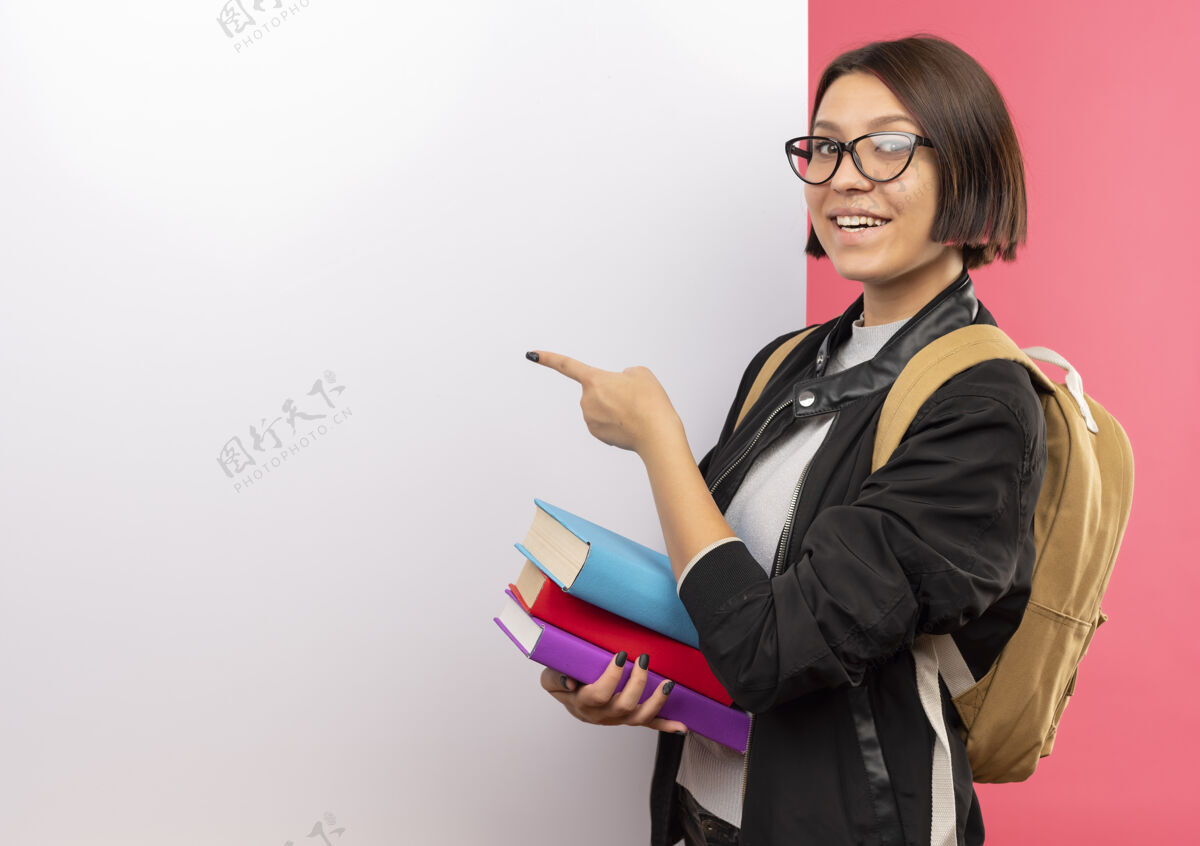 站着微笑的年轻女学生戴着眼镜 背着书包站在白色的墙前 指着隔离在粉红色墙上的墙学生穿着背