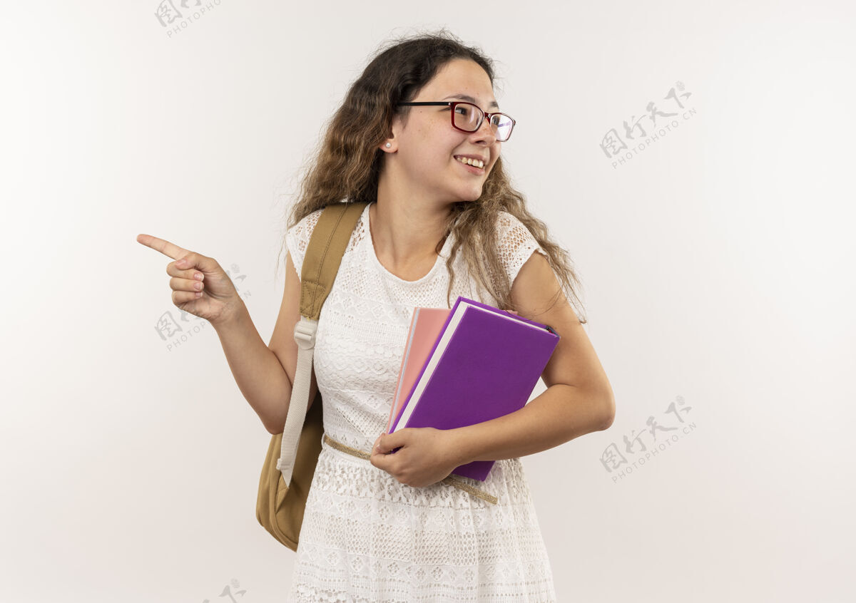 穿面带微笑的年轻漂亮的女学生戴着眼镜 背着书包 拿着笔记本 指着隔离在白墙上的一边看姿势纸条衣服