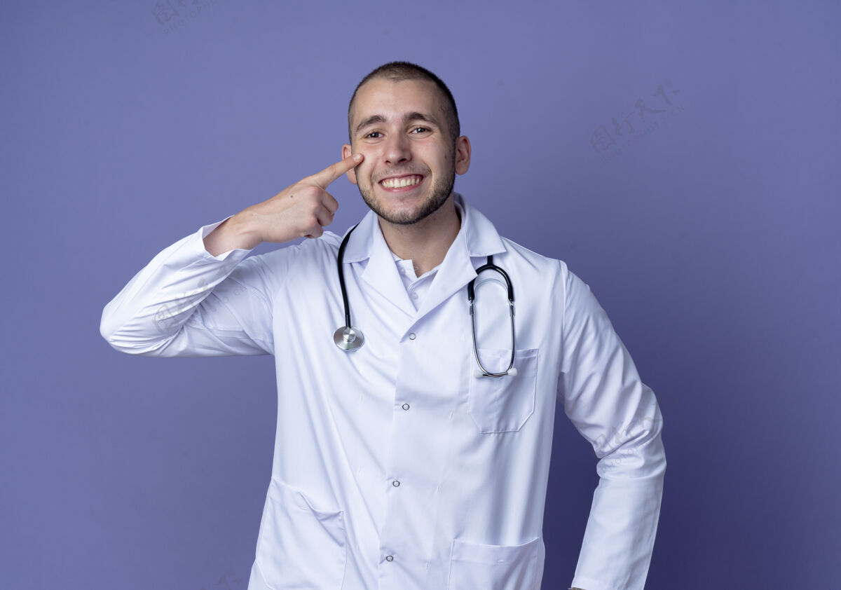 公民面带微笑的年轻男医生穿着医用长袍 听诊器把手指放在隔离在紫色墙上的脸颊上人表情人
