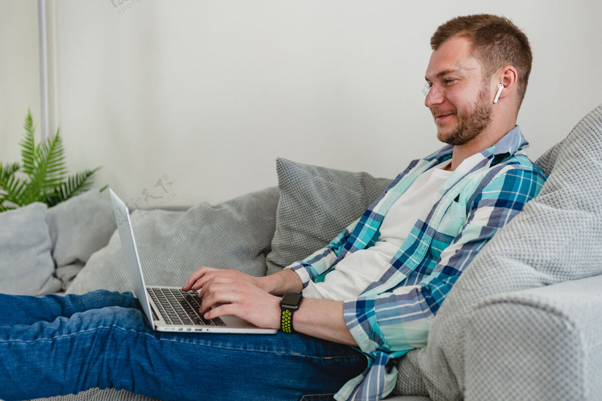 室内穿着衬衫的微笑男人在家里的沙发上放松地坐在桌子旁 在家里用笔记本电脑上网房间职业企业家