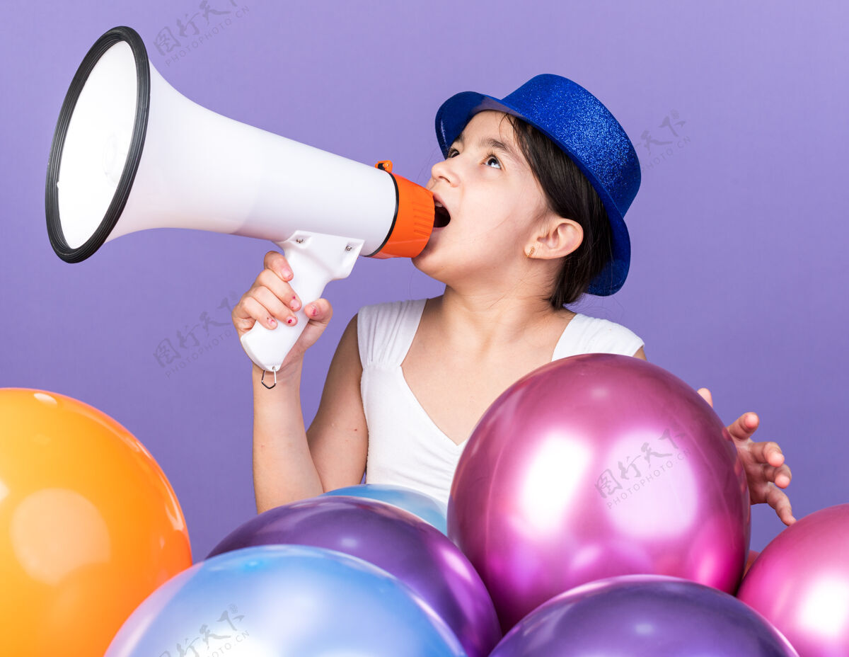 惊讶惊讶的年轻白人女孩戴着蓝色派对帽对着大声的扬声器说话 抬头看着站在紫色墙上的氦气球 上面留有复制空间大声女孩气球