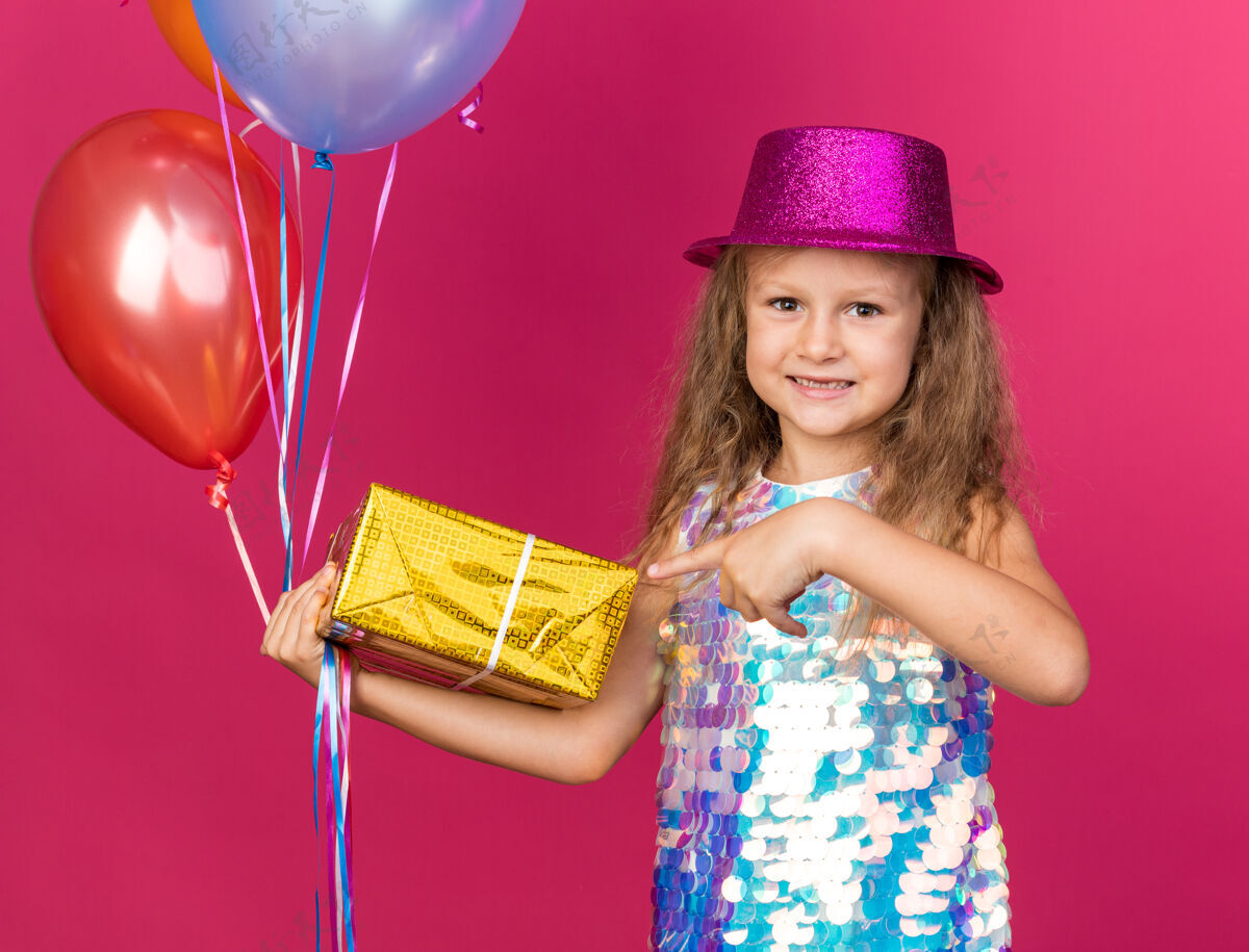 女孩微笑着的金发小女孩 戴着紫色派对帽 手里拿着氦气球 指着粉色墙壁上的礼品盒 里面有复印空间微笑生日小
