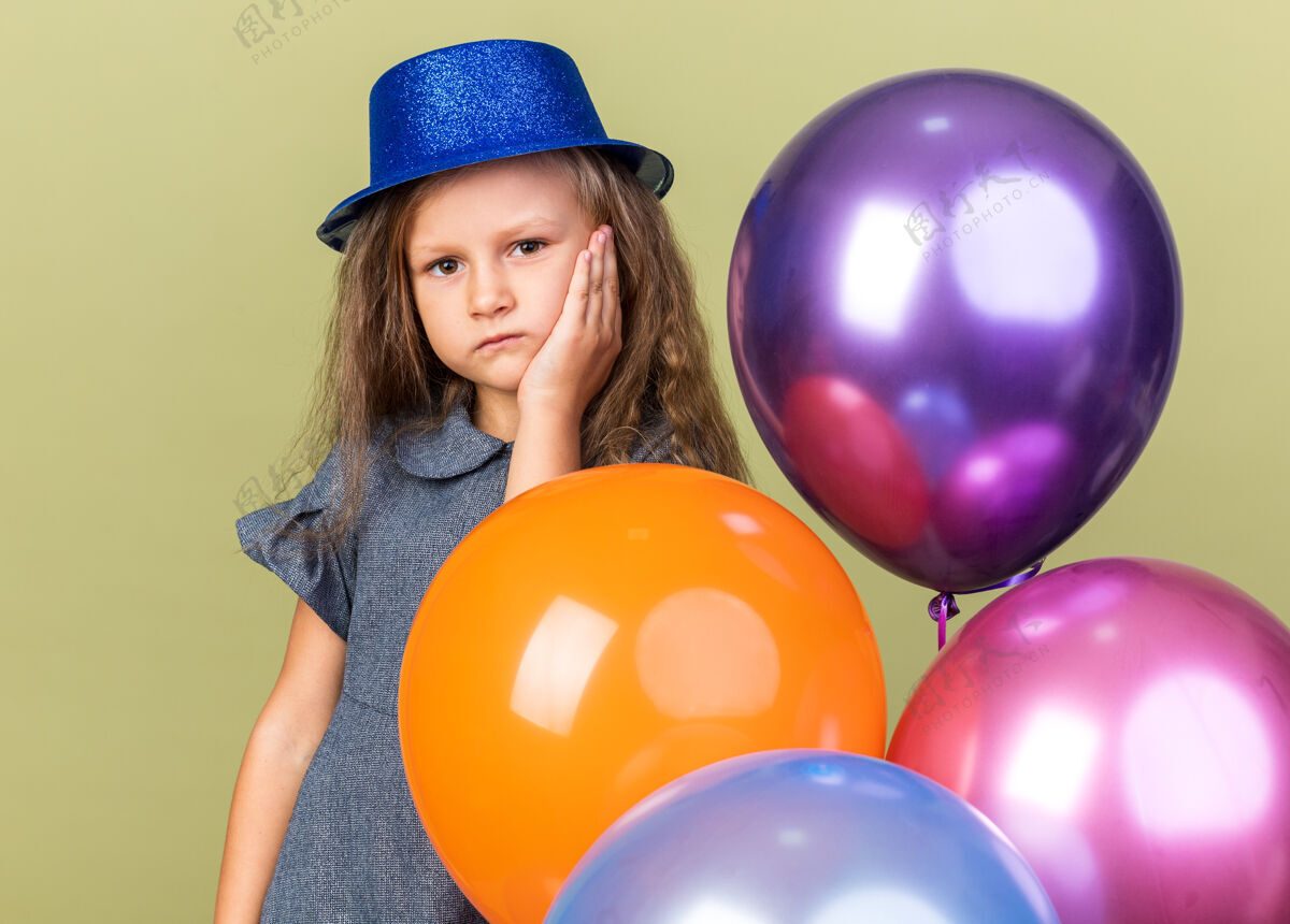 橄榄失望的金发小女孩 戴着蓝色派对帽 手放在脸上 手里拿着氦气球 孤立地放在橄榄绿的墙上 留着复制空间失望金发举行