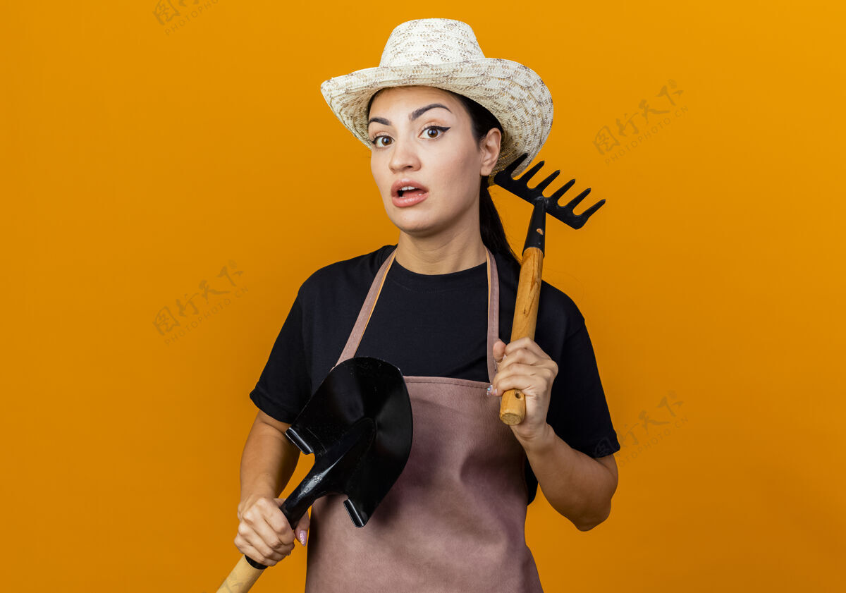 人年轻漂亮的女园丁围着围裙 戴着帽子 拿着小耙子和铲子 困惑地站在橘色的墙上看着前方迷你感觉脸