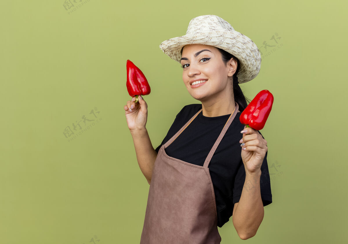 人年轻漂亮的女园丁 围着围裙 戴着帽子 手里拿着红甜椒 站在浅绿的墙上 兴高采烈地微笑着看着前面胡椒女人表情