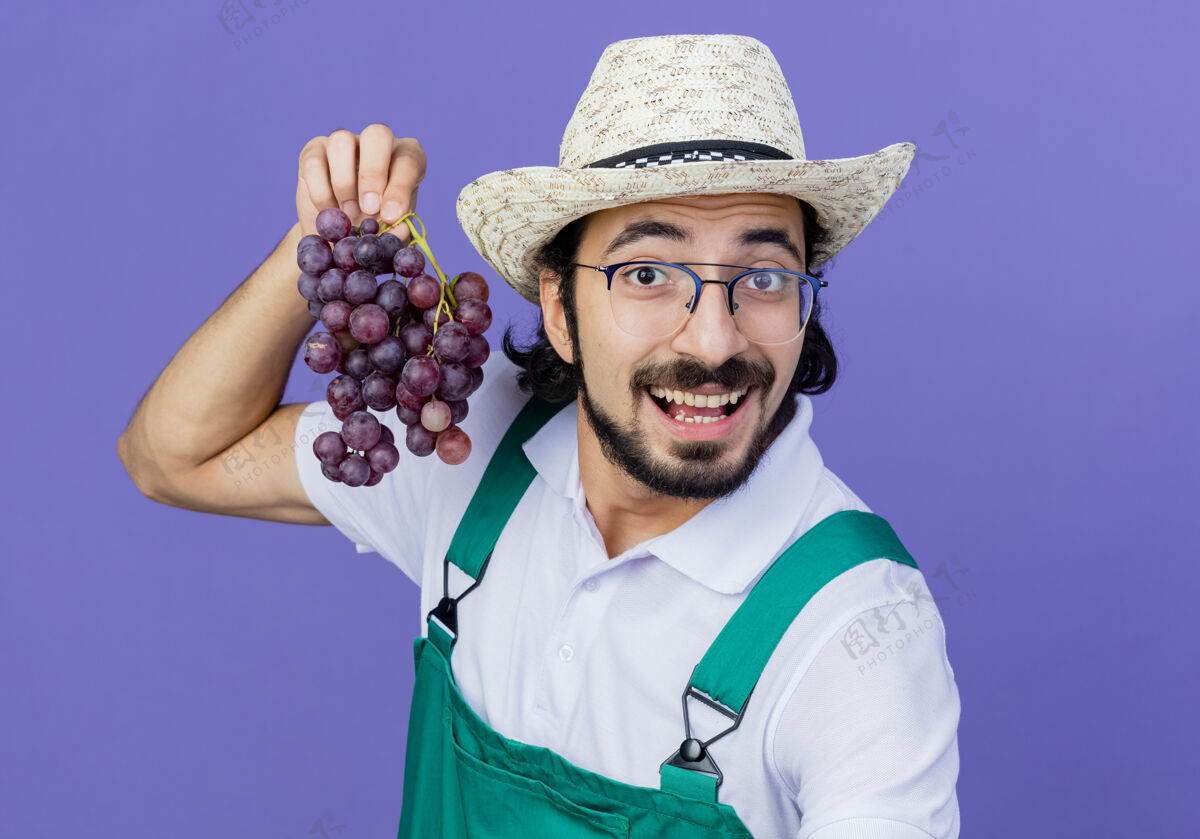 束年轻的留着胡子的园丁 穿着连体衣 戴着帽子 手里拿着一束葡萄 站在蓝色的墙上微笑着看着前面姿势衣服人