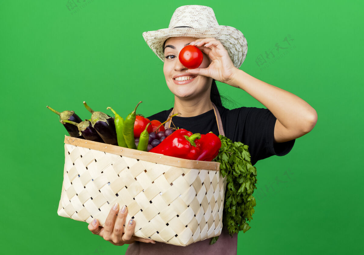 眼睛年轻漂亮的女园丁围着围裙 戴着帽子 手里拿着装满蔬菜和西红柿的篮子 站在绿色的墙上微笑着感觉篮子年轻