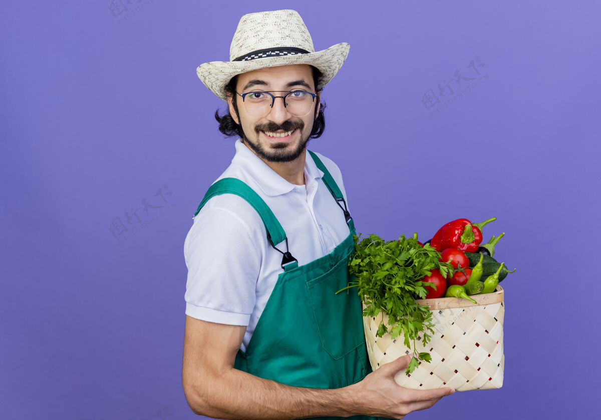 人年轻的留着胡须的园丁 穿着连体衣 戴着帽子 手里拿着装满蔬菜的箱子 站在蓝色的墙上 面带微笑地看着前方花园箱子人