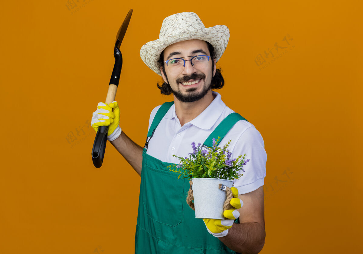 人年轻的留着胡须的园丁 穿着连体衣 戴着帽子 拿着铲子和盆栽植物 站在橙色的墙上微笑着看着前方衣服铲子连身衣