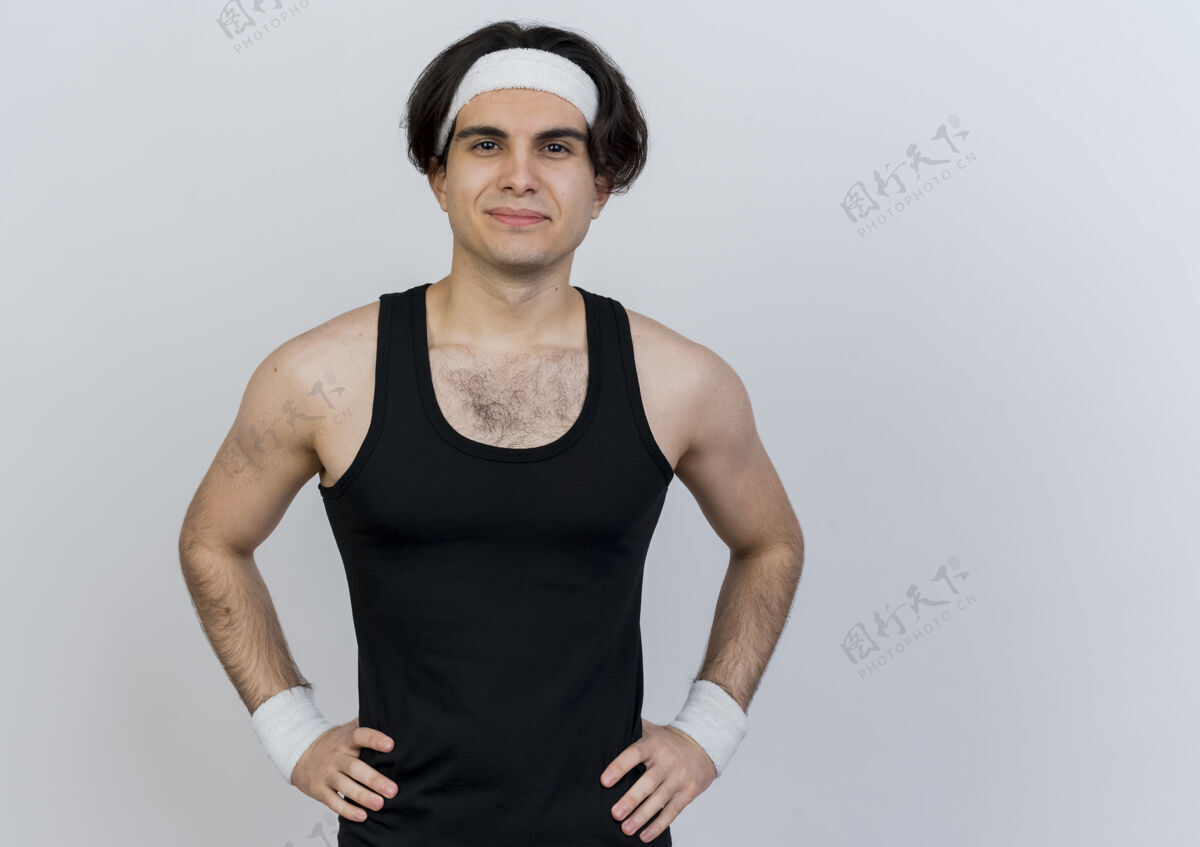 时髦年轻的运动型男子穿着运动服 戴着头巾 面带微笑 自信地站在白色的墙上 双臂放在臀部人人表情