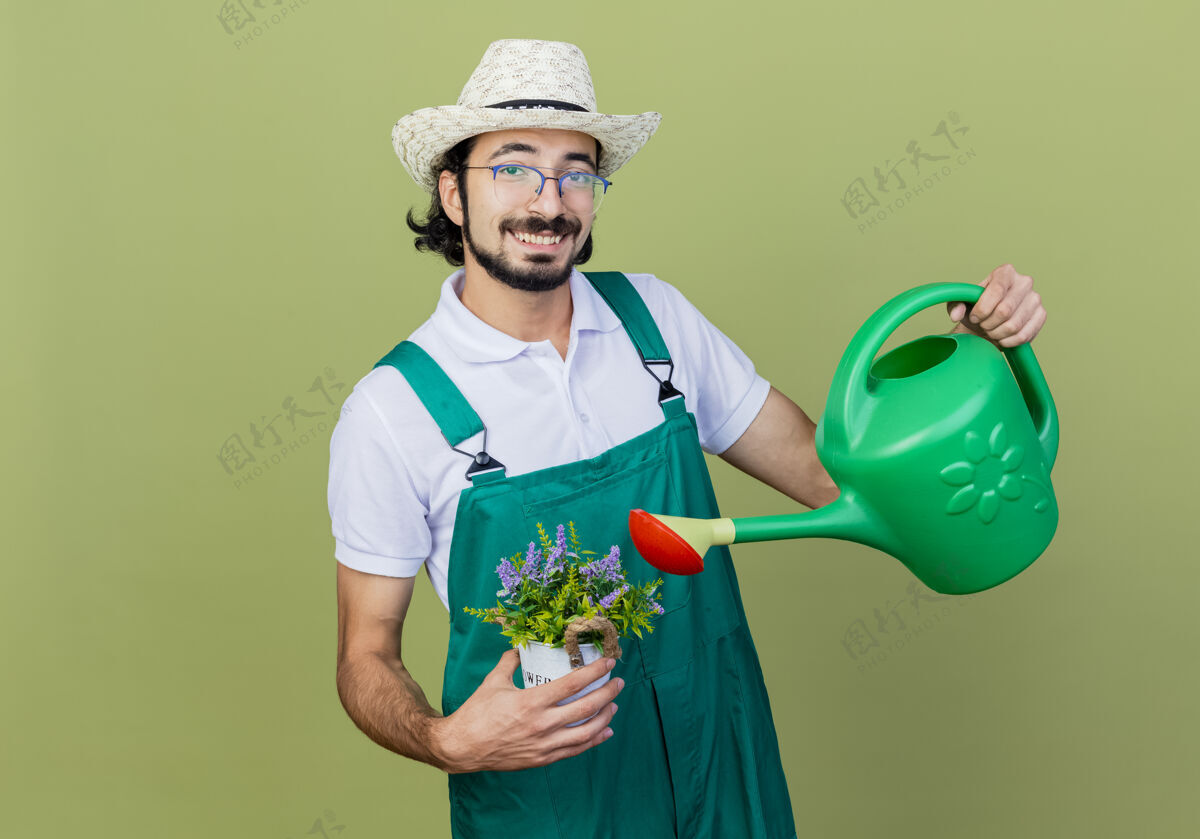 快乐年轻的留着胡须的园丁 穿着连体衣 戴着帽子 拿着水罐和盆栽植物 站在浅绿色的墙上 面带微笑地看着前方胡须穿着微笑