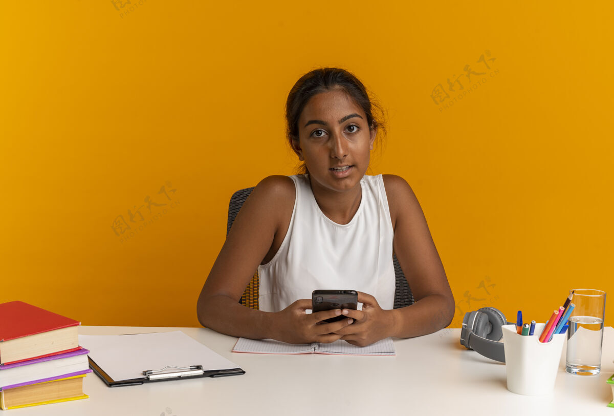 抱着看着镜头年轻的女学生坐在书桌旁拿着学习工具拿着电话坐着书桌女学生