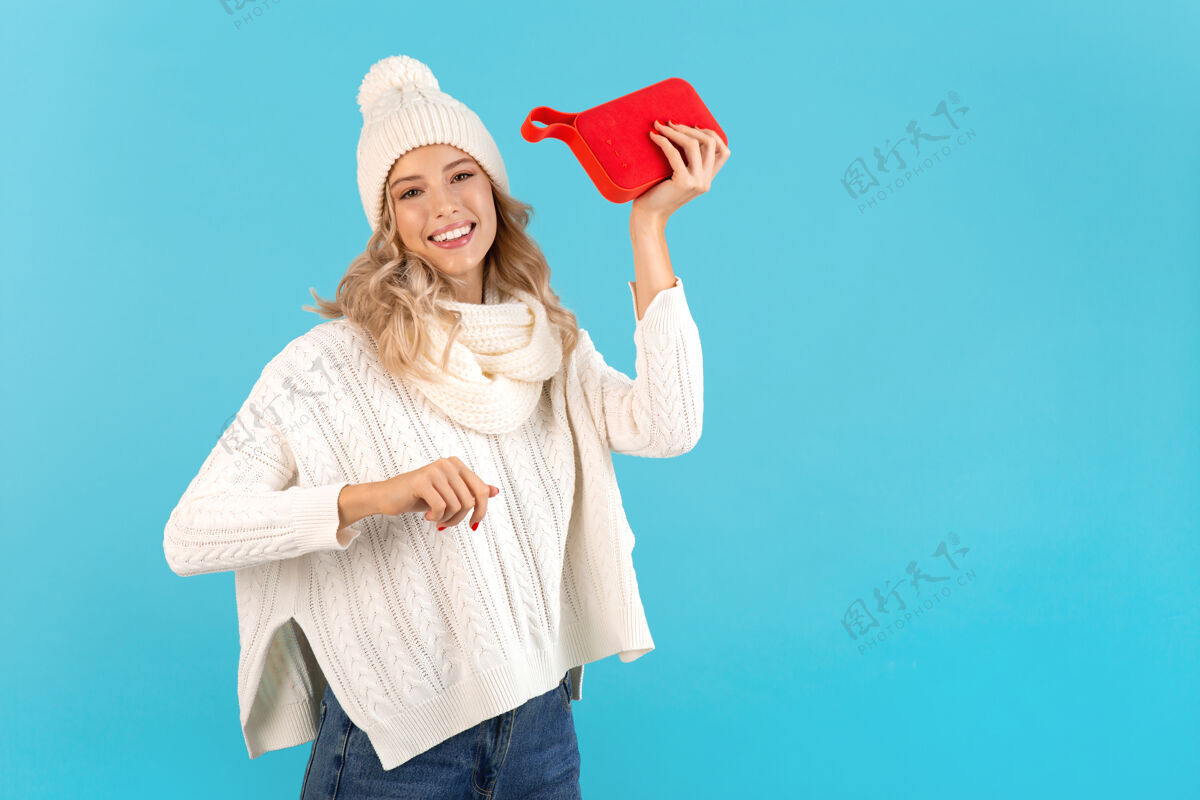 乐趣时尚的金发微笑的美丽年轻女子手持无线扬声器听音乐穿着白色毛衣和针织帽子摆在蓝色配件欢呼红色