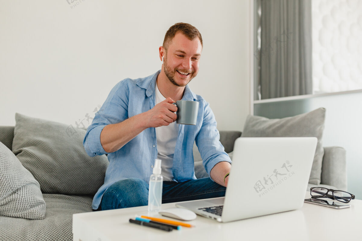 休闲微笑的男人坐在沙发上喝茶 在家里的桌子上用笔记本电脑上网职业使用笔记本电脑