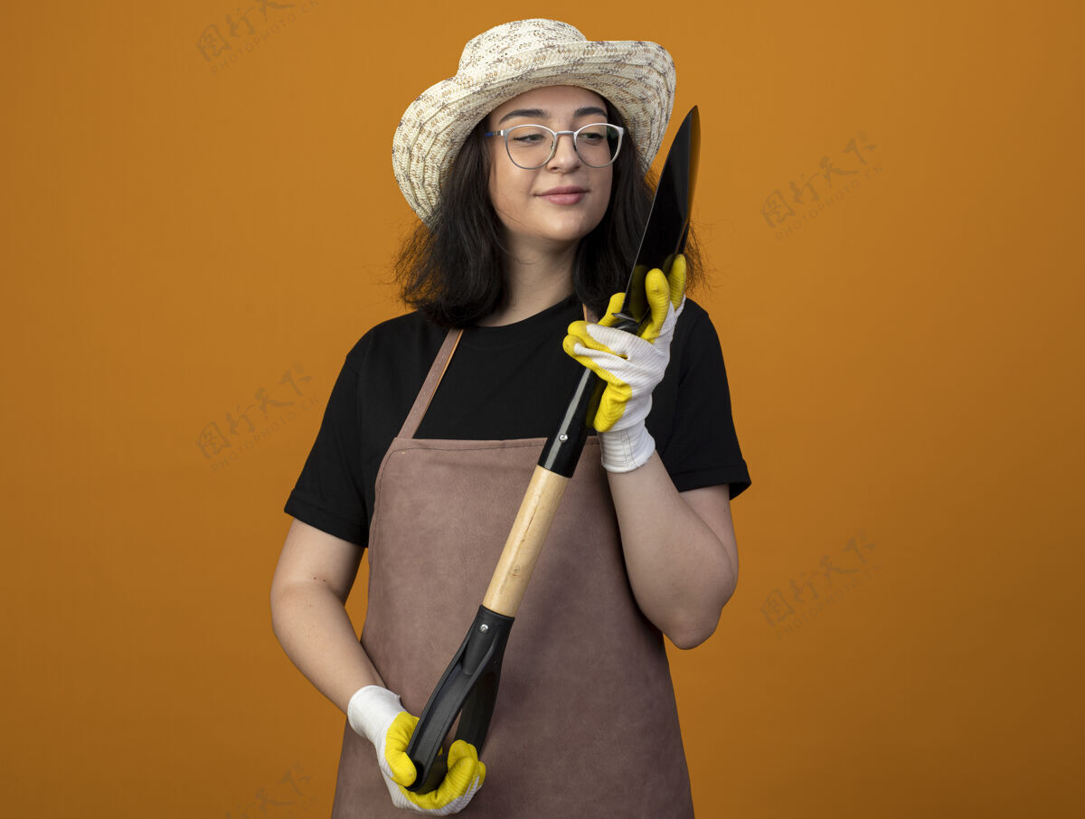 女人高兴的是 年轻的黑发女园丁戴着眼镜 穿着制服 戴着园艺帽和手套 手里拿着一把铲子 看着隔离在橙色墙上的铲子市民制服帽子