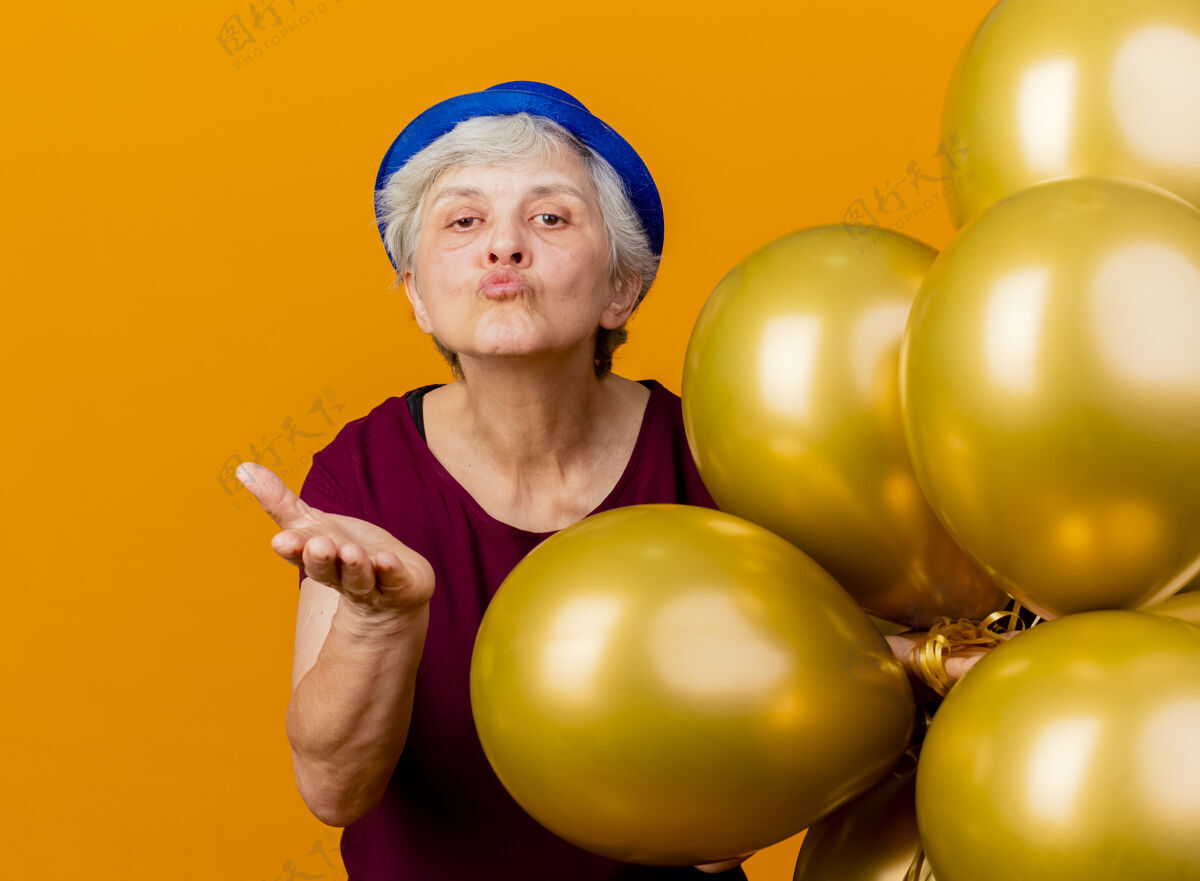 女人戴着派对帽的自信的老妇人手持氦气球 用隔离在橙色墙上的手送上吻氦聚会姿势