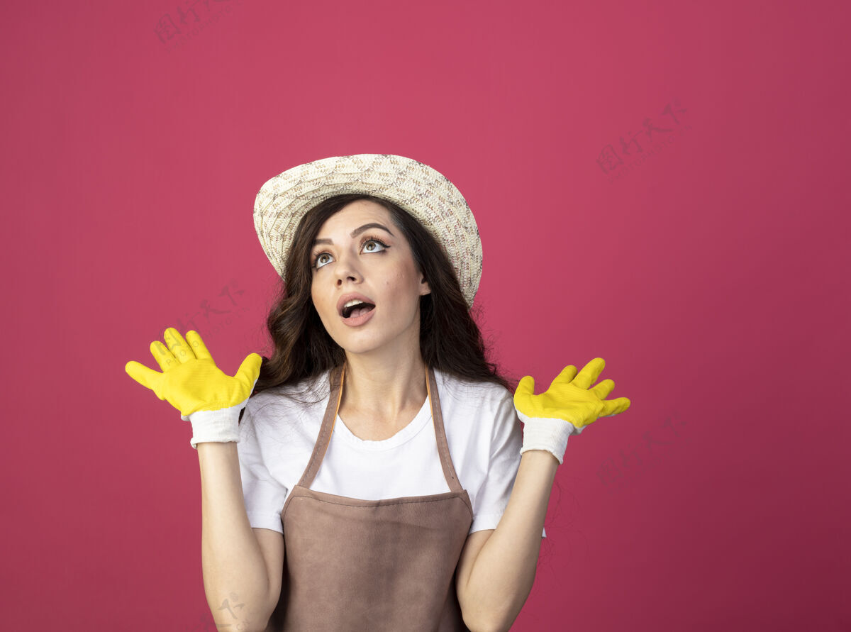 制服穿着制服 戴着园艺帽和手套的年轻女园丁惊讶地站在粉红色的墙上 抬起双手 孤零零地看着手年轻人女人