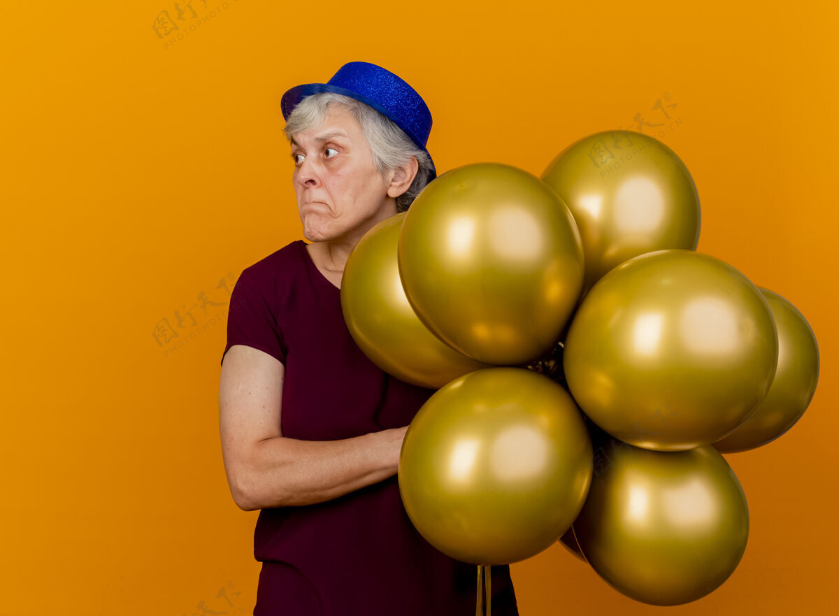 脸戴着派对帽的焦虑的老妇人拿着氦气球 看着橙色墙上孤立的一面姿势焦虑老人
