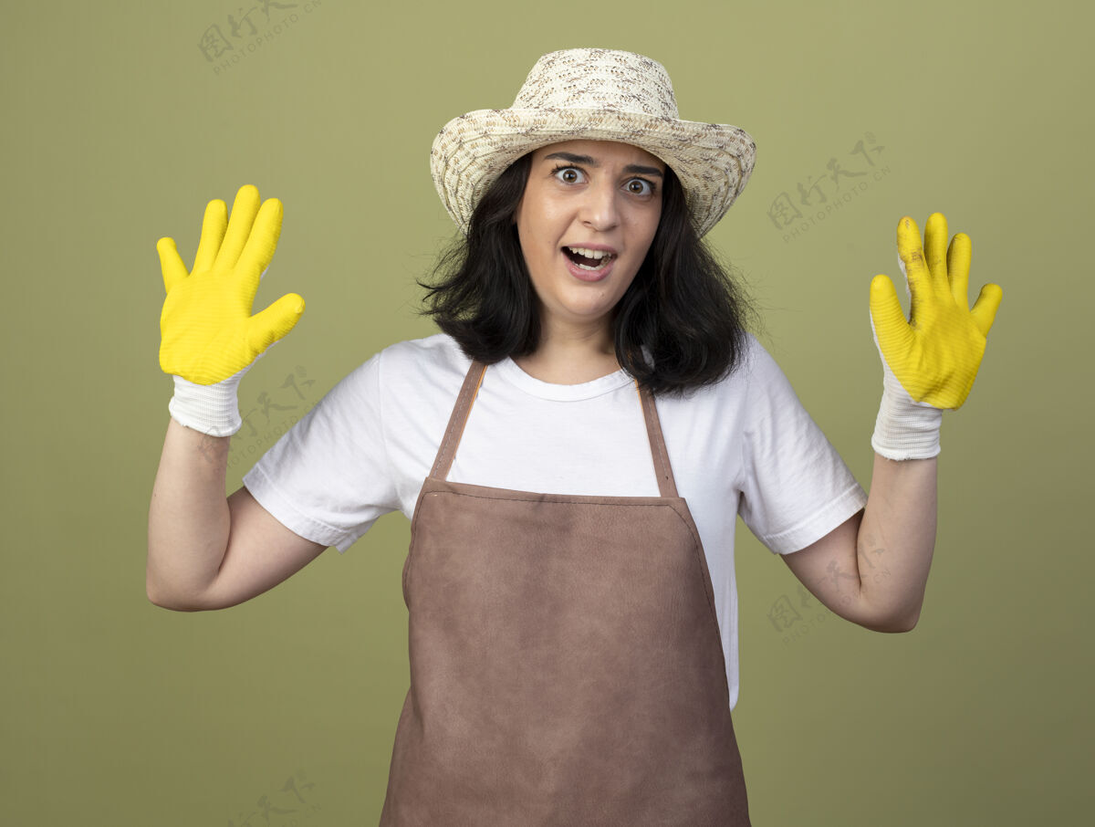 手套焦急的年轻黑发女园丁穿着制服戴着园艺帽和手套站在橄榄绿的墙上 举手孤立制服市民人
