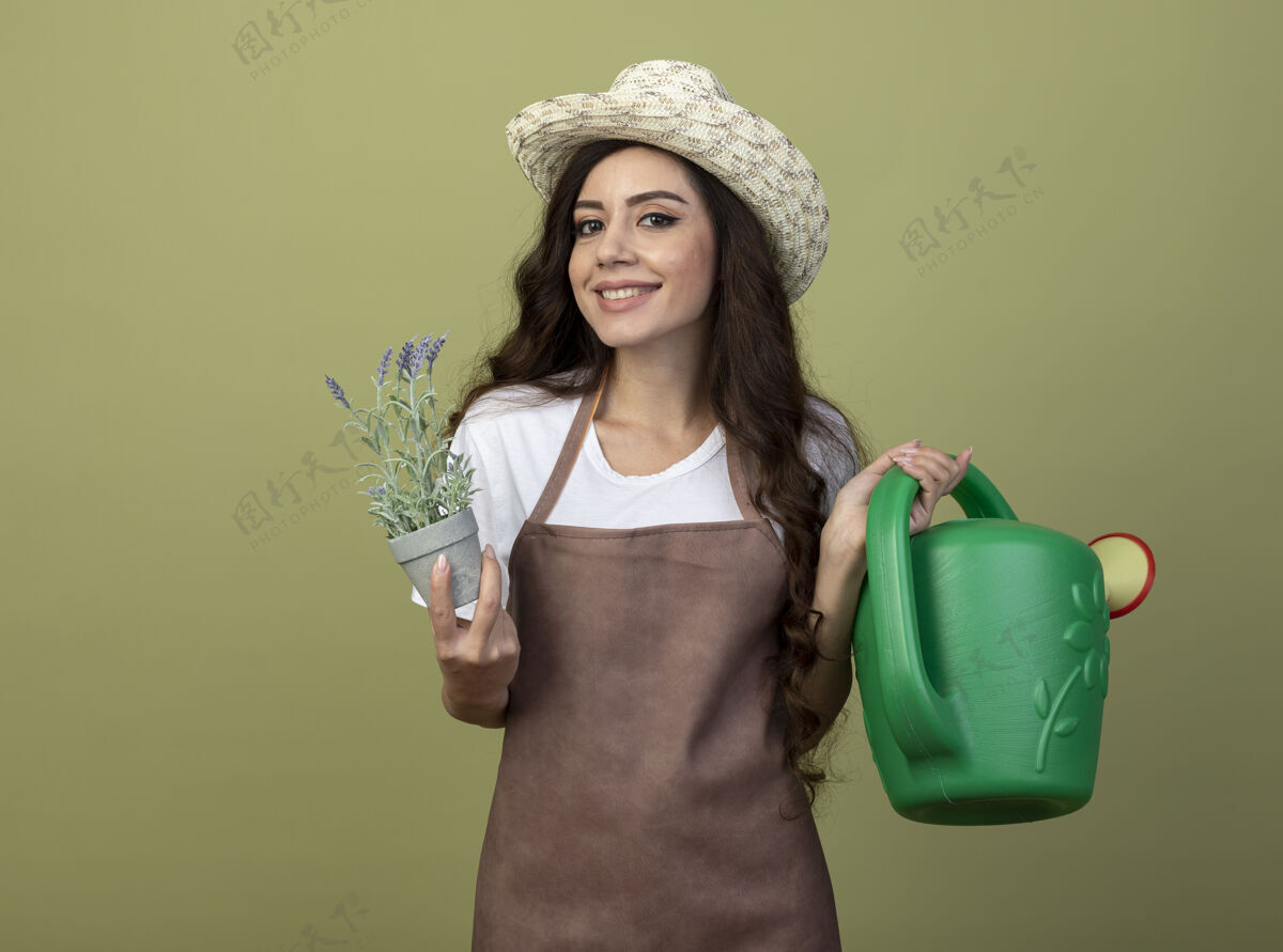 举行微笑着的年轻女园丁穿着制服 戴着园艺帽 手持花盆和浇水罐 隔离在橄榄绿的墙上人制服穿着