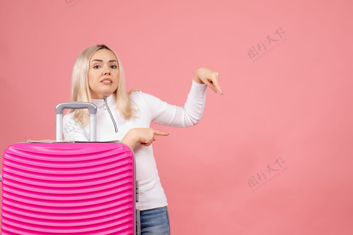 成人前视图：站在粉色手提箱后面的年轻女士展示了一些东西欢乐风景小姐