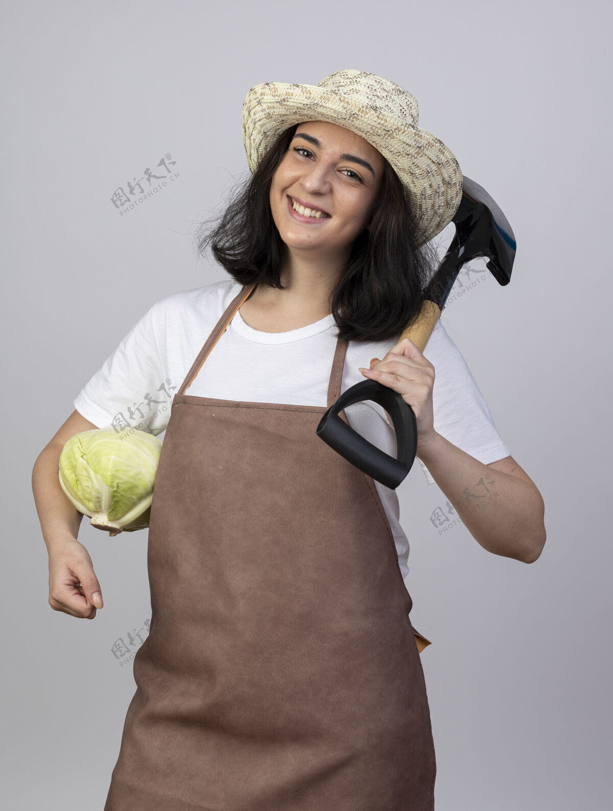 微笑微笑的年轻黑发女园丁穿着制服 戴着园艺帽 肩上扛着卷心菜和铲子 隔离在白墙上园艺花园黑发