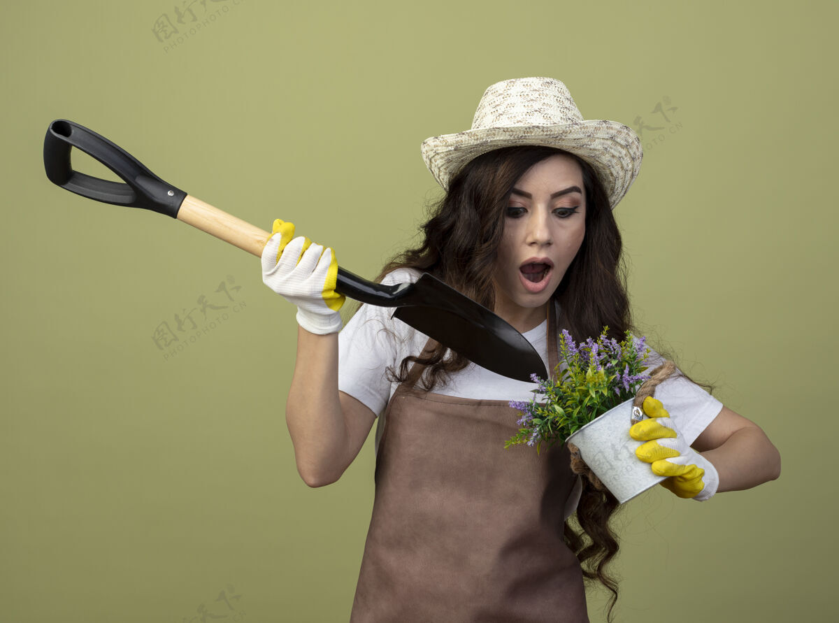花园穿着制服 戴着园艺帽和手套的年轻女园丁惊讶地把铁锹举过橄榄绿墙上孤立的花盆黑桃人帽子