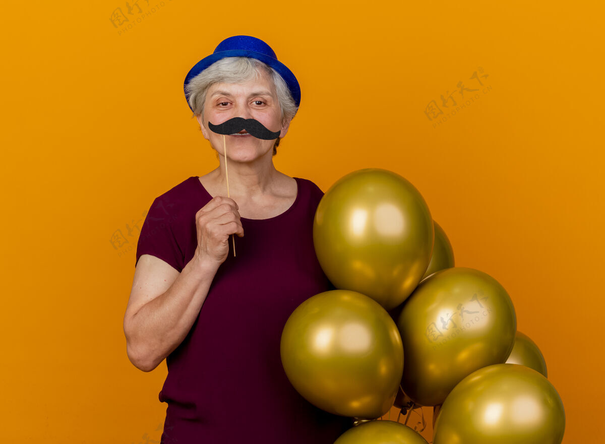 人微笑着戴着派对帽的老妇人拿着氦气球和假胡子在橘色墙上孤立的棍子上老人胡子表情