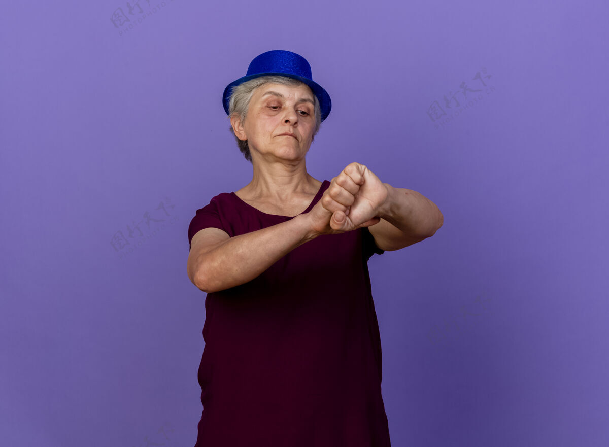表情戴着派对帽的自信的老妇人抱着 看着隔离在紫色墙上的手臂人公民手臂