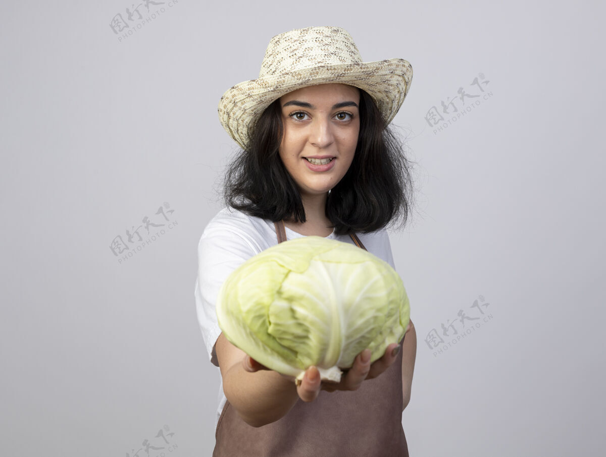 女性微笑着的年轻黑发女园丁穿着制服 戴着园艺帽 把卷心菜隔离在白墙上人年轻人脸