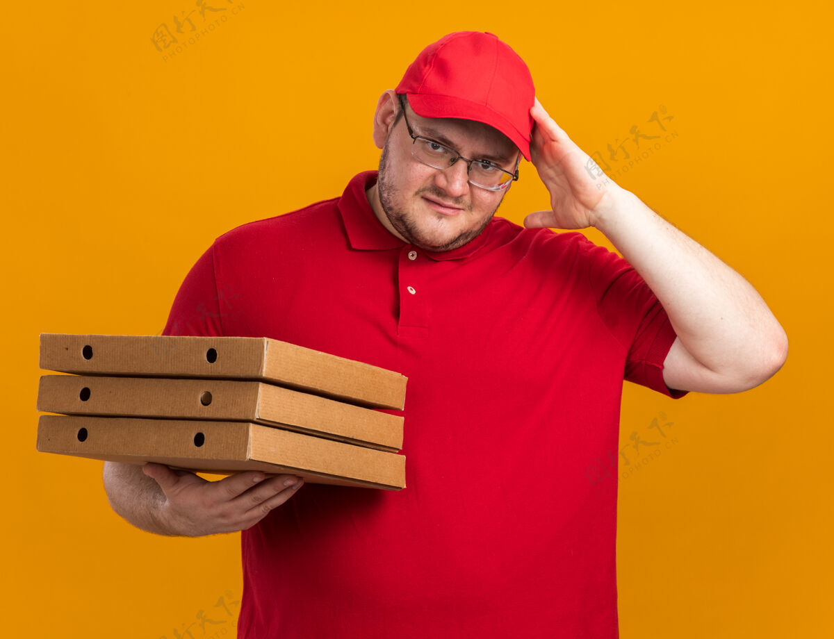 困惑困惑超重的年轻送货员戴着眼镜拿着披萨盒 手放在头上 隔离在橙色的墙上 留着复印空间持有眼镜年轻