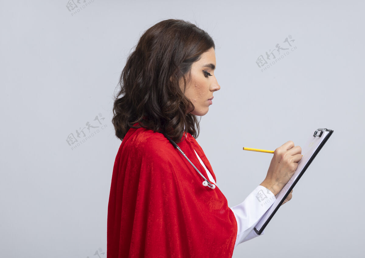 女孩自信的女超人穿着医生制服 穿着红色斗篷和听诊器 站在一边 拿着剪贴板和铅笔 隔离在白色的墙上制服拿着站立